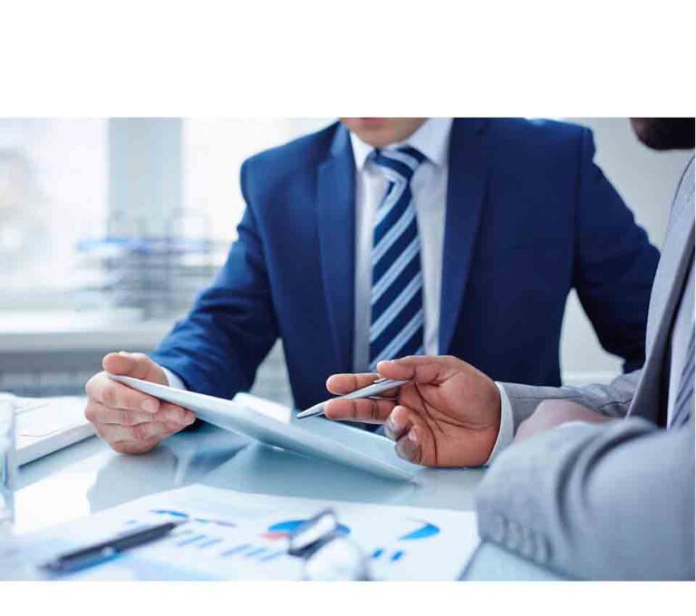 Two people in suits discuss business documents at a table, one holding a pen and the other a tablet.