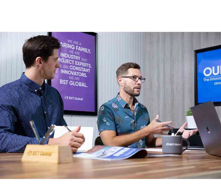 Two men sit at a table, engaged in discussion. One gestures while speaking. A laptop, notepad, and coffee mugs are on the table. Posters with text are on the wall behind them.