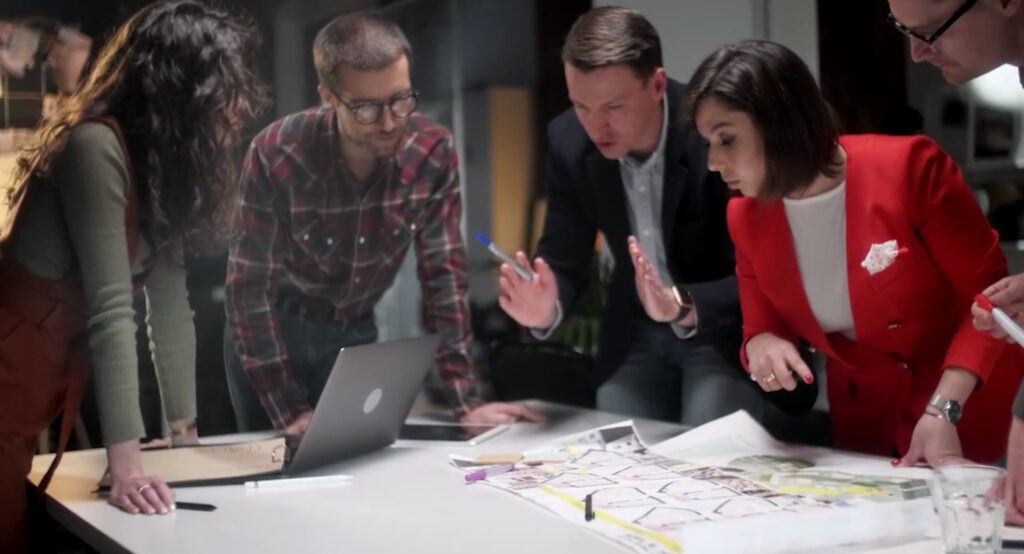 Five people gathered around a table, looking at a laptop and discussing a printed layout.