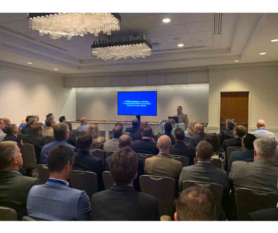 A full room of people seated, facing a speaker at a podium. A presentation slide is displayed on a screen behind the speaker.