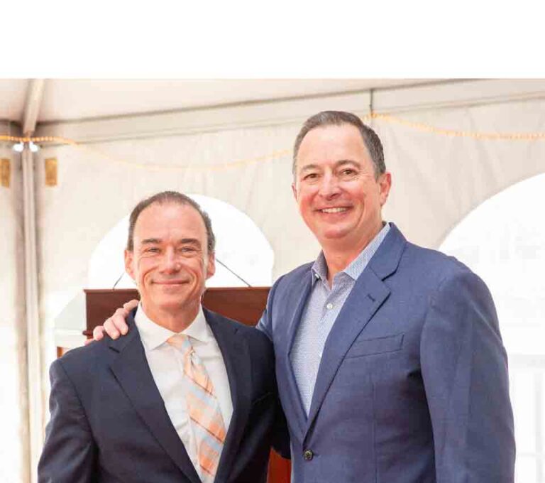 Two men in suits smiling, one has an arm around the other's shoulder, standing under a tent.