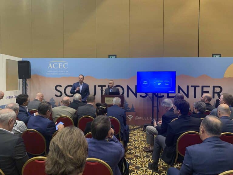 Two speakers present at a conference in front of an audience. The backdrop reads "Solutions Center" with a logo for ACEC.