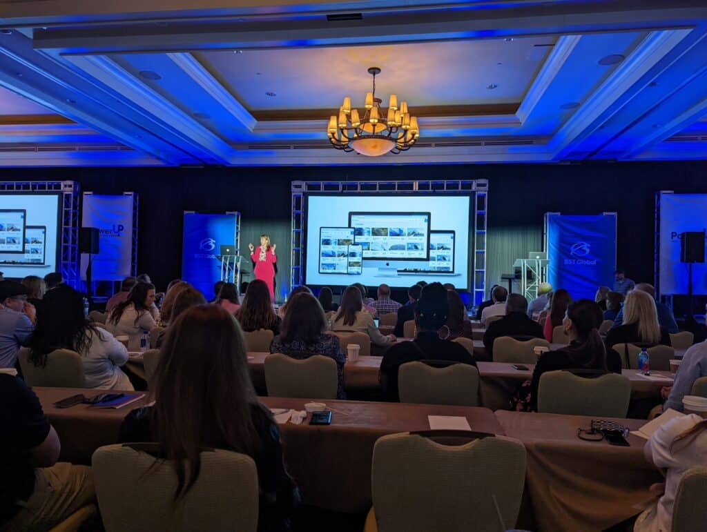 Audience seated at a conference, facing a large screen displaying slides and two presenters on stage.