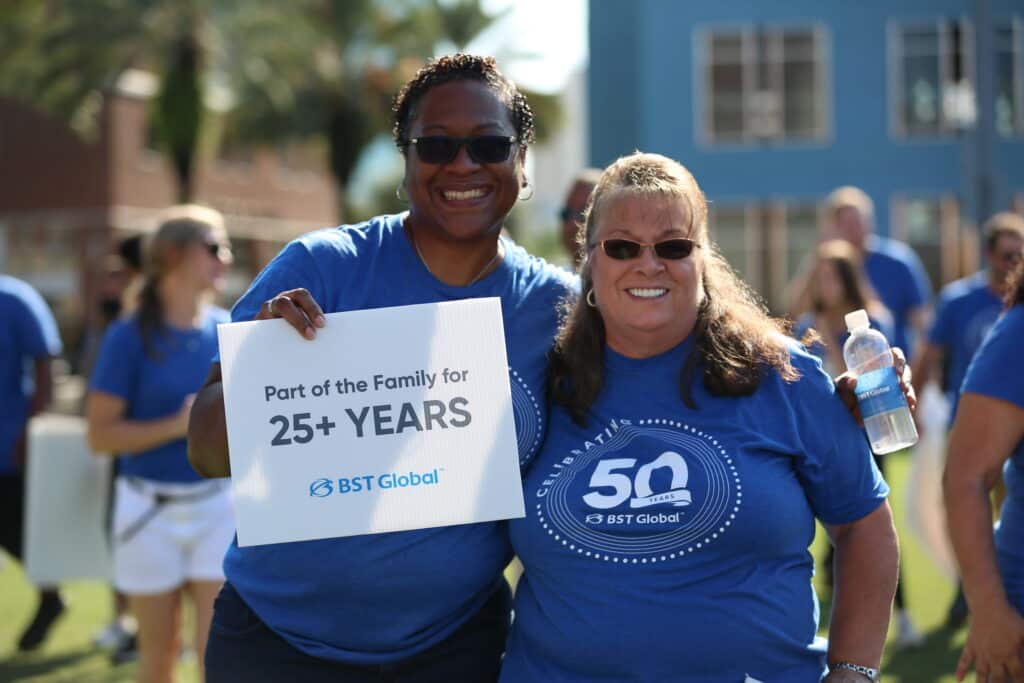 Two people smiling at a celebration event, one holding a sign that reads "Part of the Family for 25+ Years," with others in blue BST Global shirts in the background.
