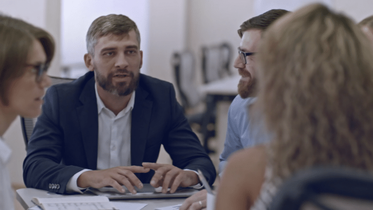 A group of people in a meeting room, engaged in discussion around a table with documents.
