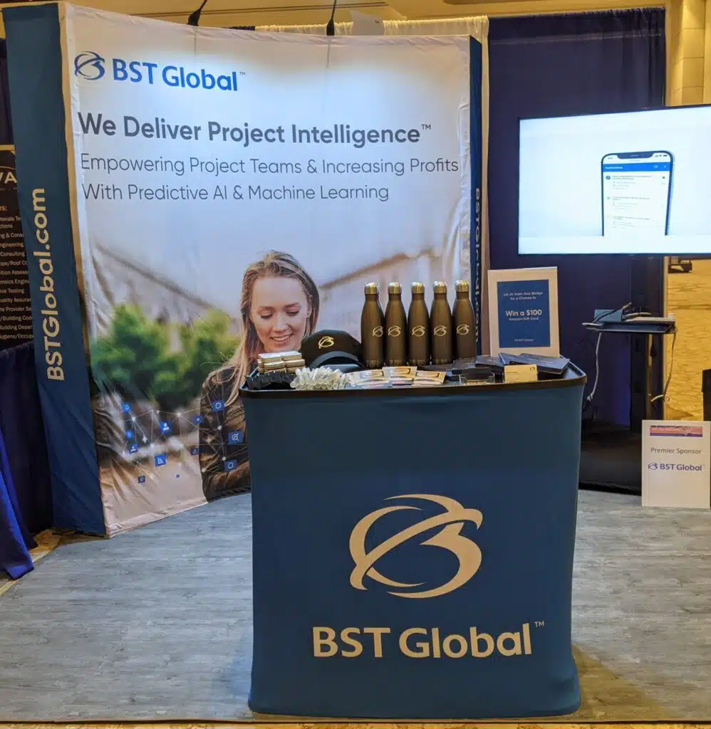 A trade show booth for BST Global featuring a sign with a smiling person, company branding, a table with merchandise, and promotional materials.