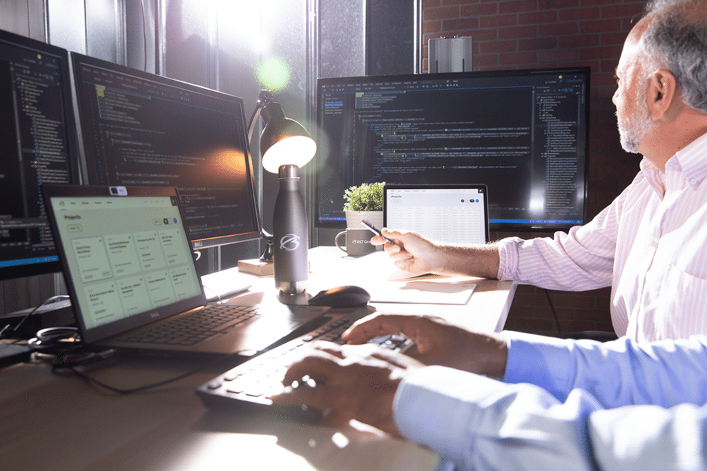 Two people working at a desk with multiple computer screens displaying code and project management software, in a well-lit office space.