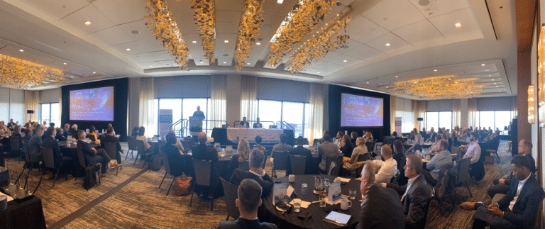 A conference room with people seated, facing a stage with a panel and speakers. Large screens display presentations. Overhead decorative lights are visible.