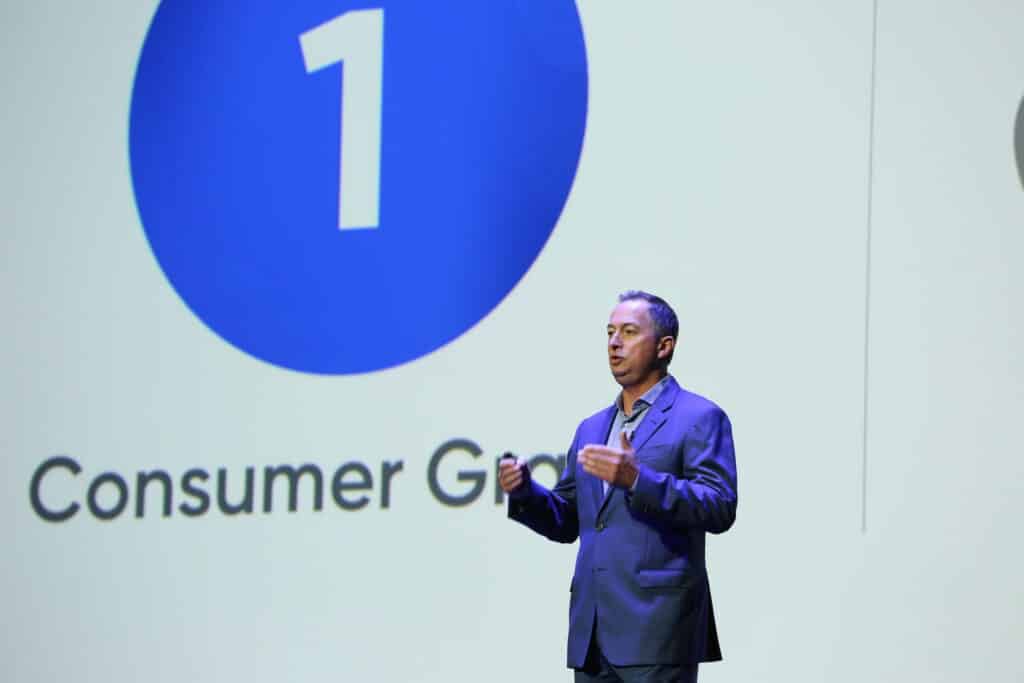 Man in a suit giving a presentation on stage. Large blue circle with the number "1" and the text "Consumer Goal" displayed on a screen behind him.