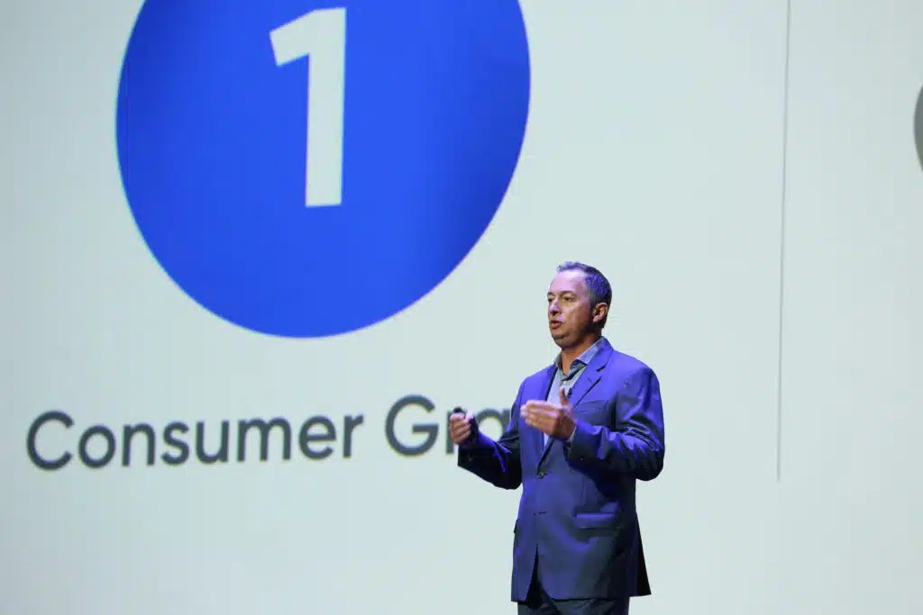 Man in a suit giving a presentation on stage. Large blue circle with the number "1" and the text "Consumer Goal" displayed on a screen behind him.