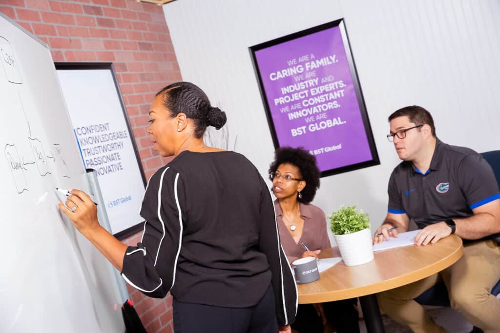Three people in a meeting room, one writing on a whiteboard while two others sit at a round table discussing. A poster about company values is on the wall.
