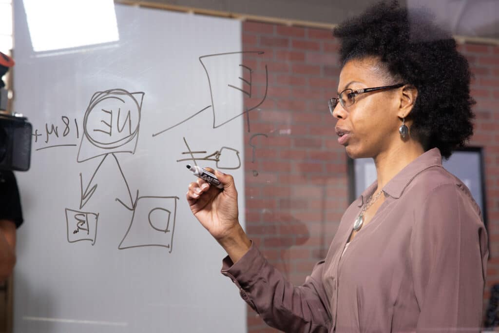 A person writes diagrams and notes on a transparent board with a marker.