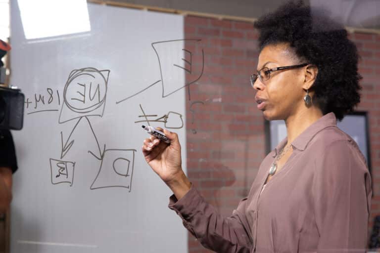 A person writes diagrams and notes on a transparent board with a marker.