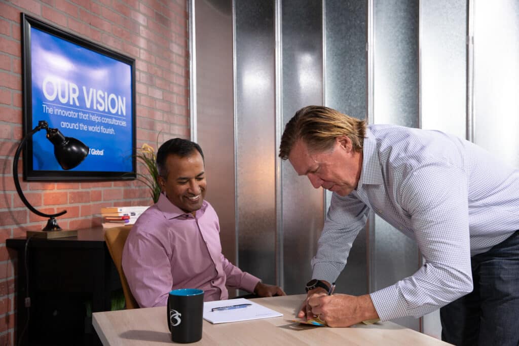 Two men in an office, one sitting and smiling, the other signing a document on a desk. A screen displays a vision statement.