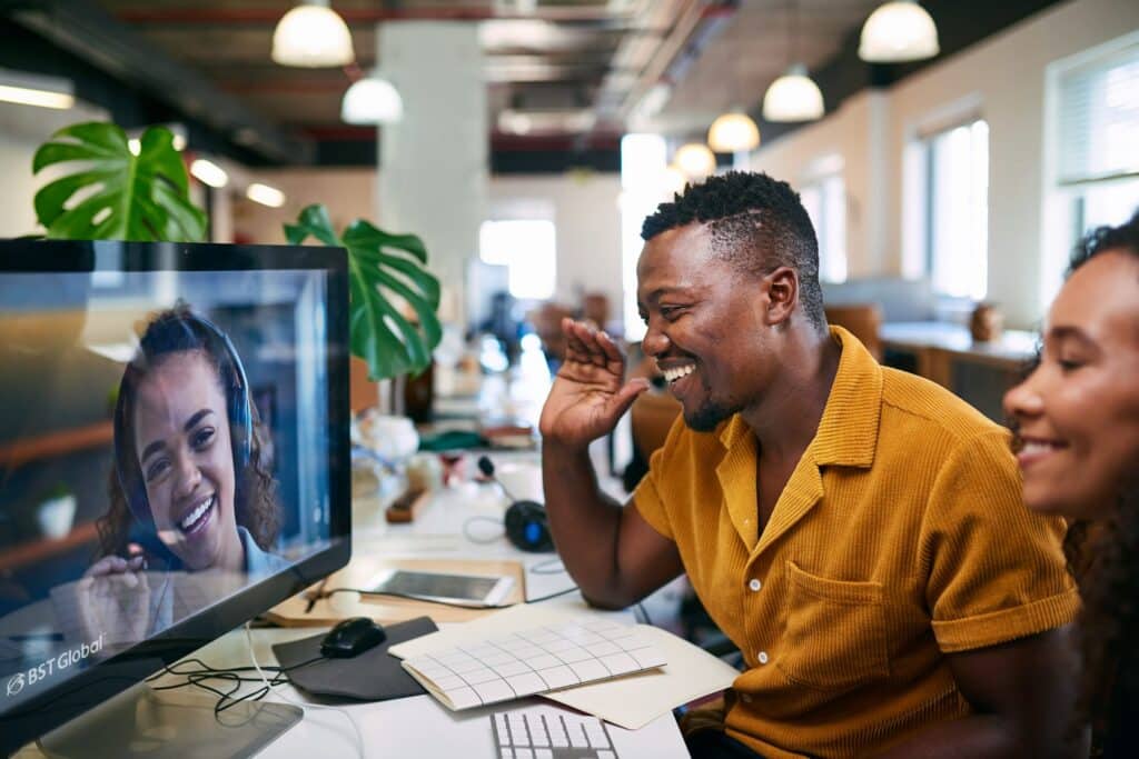 A man waves at a woman on a video call displayed on a computer screen in a modern office setting.