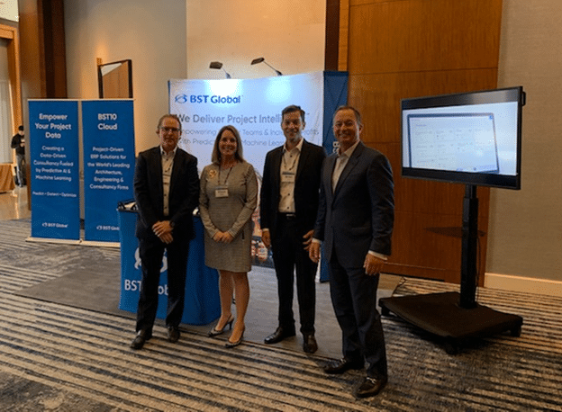 Four people stand in front of a BST Global display booth at an event. A screen to the right shows a presentation slide.