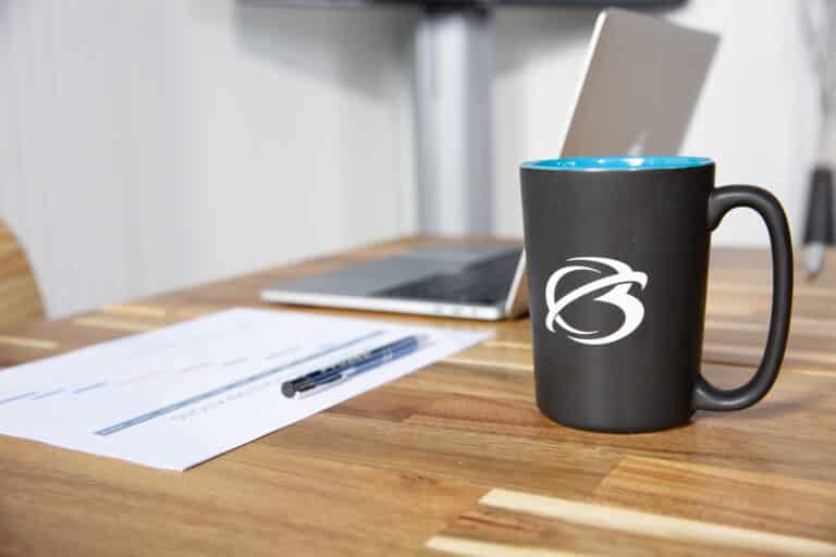 A black mug with a logo sits on a wooden table next to a pen, paper, and an open laptop.