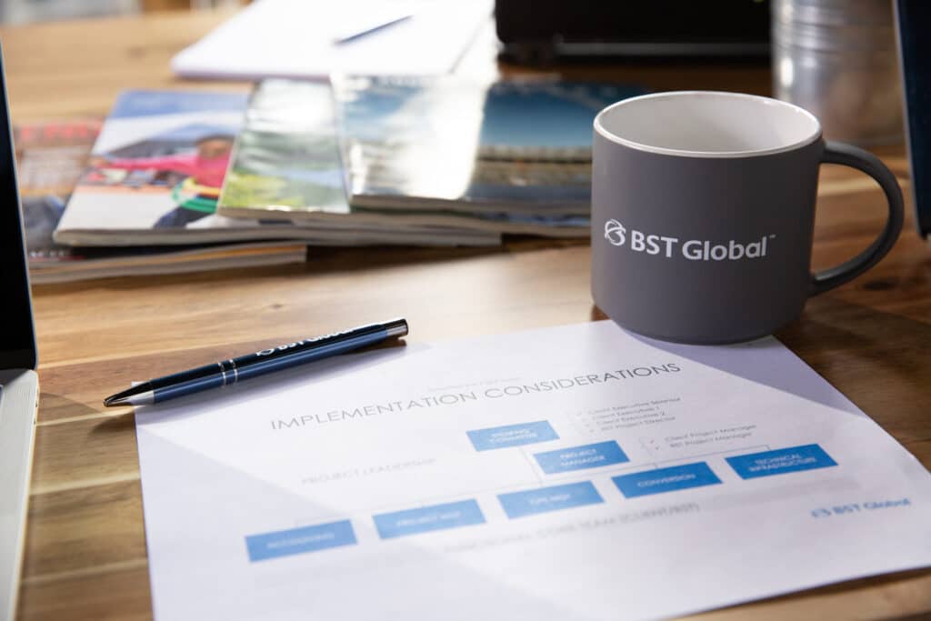 A mug labeled "BST Global" sits on a desk with a document titled "Implementation Considerations," a pen, and some magazines.