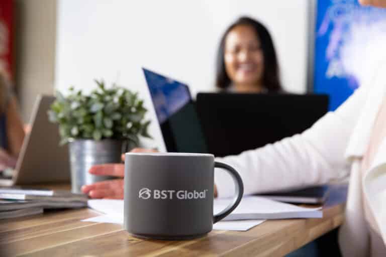 A gray mug with "BST Global" logo is on a wooden table, alongside papers and a potted plant. A person is blurred in the background, working on a laptop.