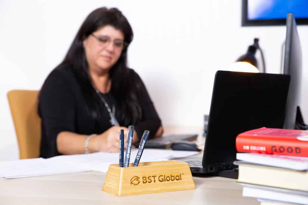 Woman working at desk with a laptop, pens in a BST Global holder, and books on the side.