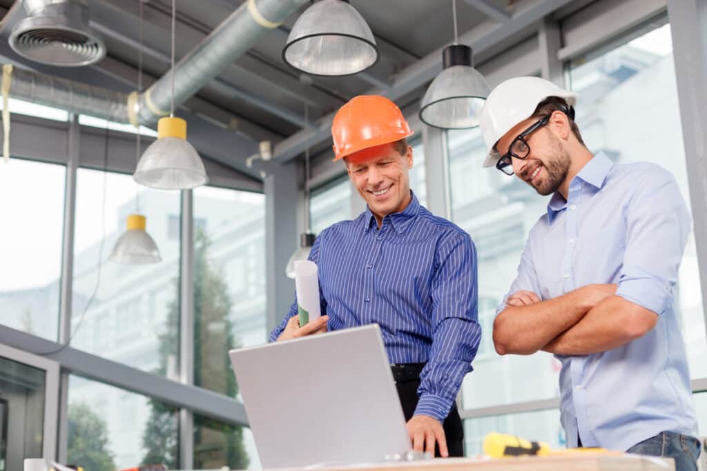 Two men wearing hard hats review a project on a laptop in a modern office with large windows and industrial lighting.