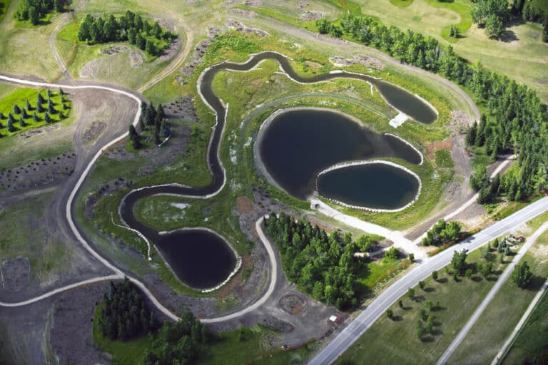 Aerial view of a landscaped park area with ponds, winding paths, and surrounding greenery. A road borders the park on the right.