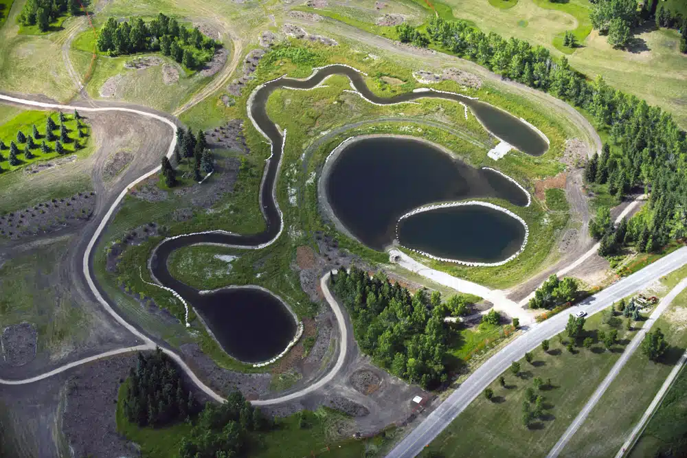 Aerial view of a landscaped park area with ponds, winding paths, and surrounding greenery. A road borders the park on the right.