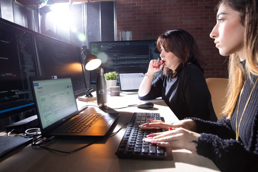 Two people work at a desk with computers and keyboards, focused on programming tasks in a modern office setting.