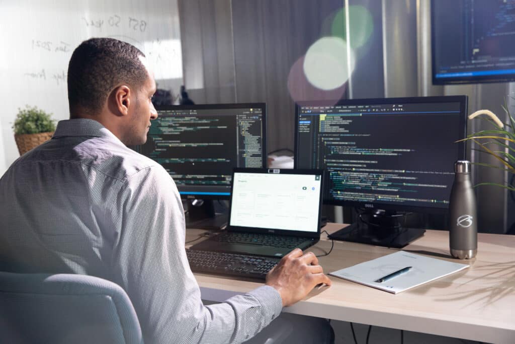 A person working at a desk with three computer monitors displaying code, a laptop, a tablet, and a water bottle nearby.