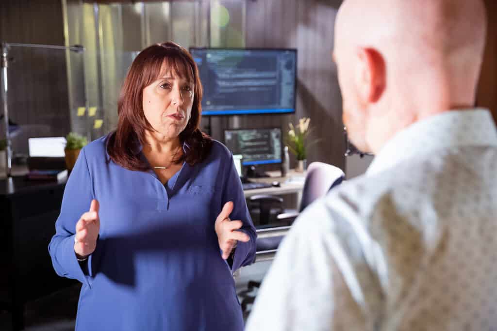 Two people are talking in an office with computer monitors in the background. One person gestures with hands while the other listens.