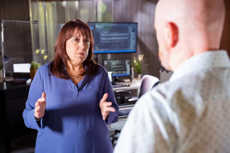 Two people are talking in an office with computer monitors in the background. One person gestures with hands while the other listens.
