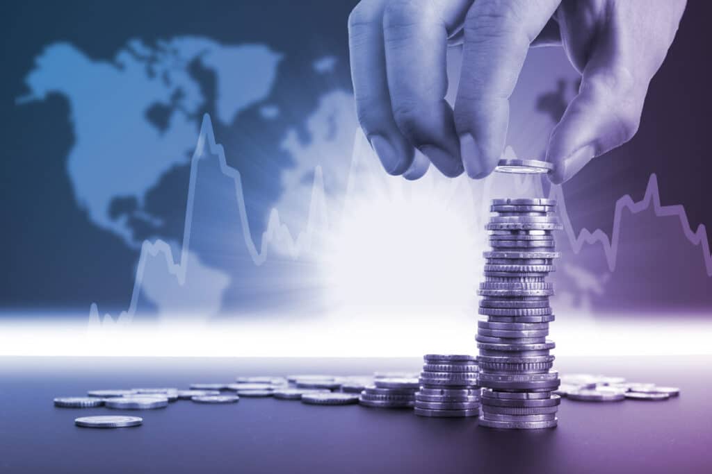 A hand stacks coins in front of a digital world map with fluctuating graph lines, symbolizing global finance or economic growth.