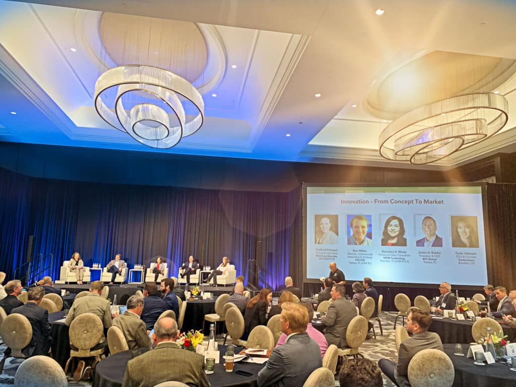A panel of speakers on stage at a conference with attendees seated at round tables. A large screen displays speaker information.