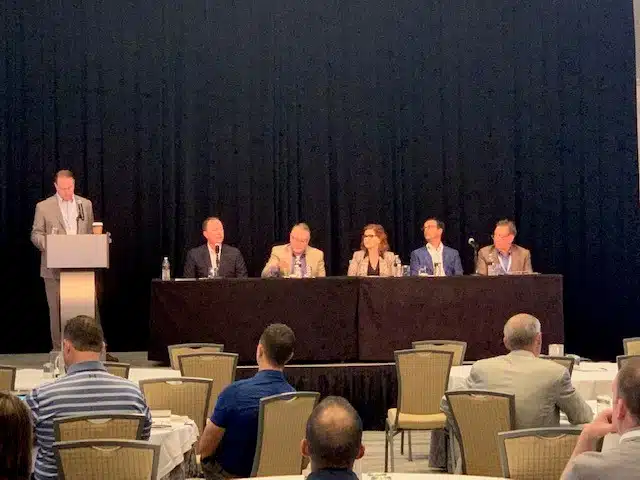 A panel of six people on stage with one person standing at a podium and five seated at a table, speaking to an audience in a conference setting.