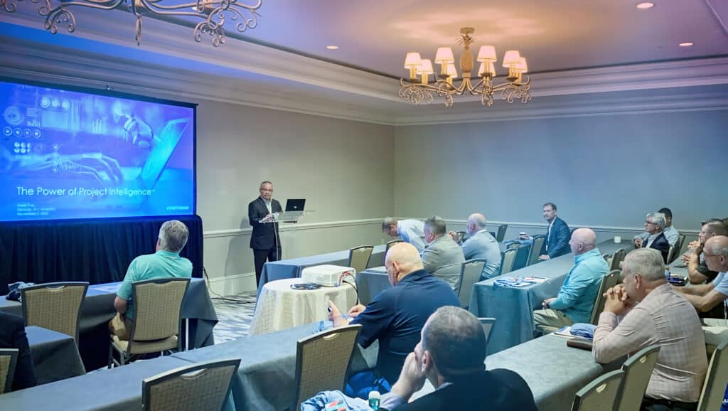 A speaker presents a slideshow titled "The Power of Project Intelligence" to an audience in a conference room with chandeliers.