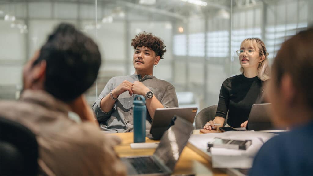 A group of people sitting at a conference table with laptops and documents, engaged in a discussion in an office setting.