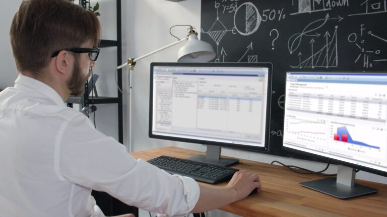 A person works at a desk with dual monitors displaying data and charts, in front of a chalkboard with mathematical diagrams and graphs.