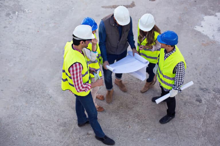 A group of construction workers wearing hard hats and reflective vests stand in a circle examining blueprints.