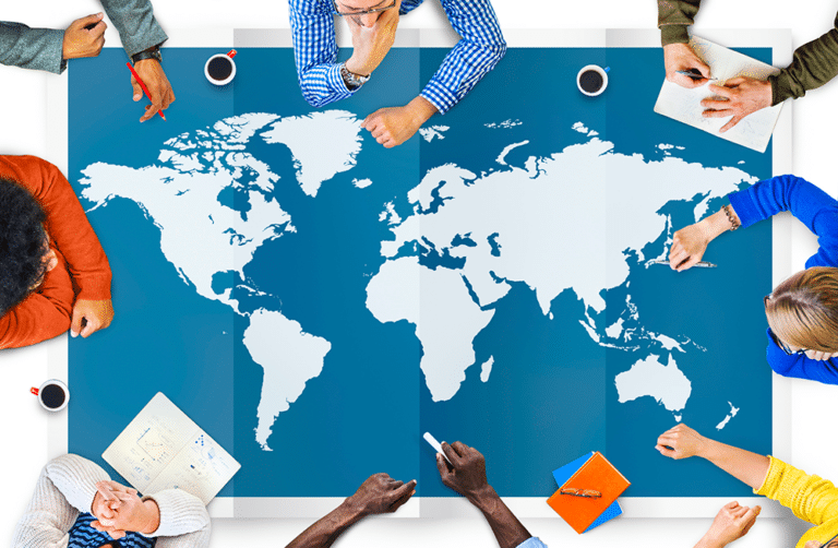 A group of diverse individuals sit around a table with a world map, pointing and discussing. Various notebooks and coffee cups are on the table.