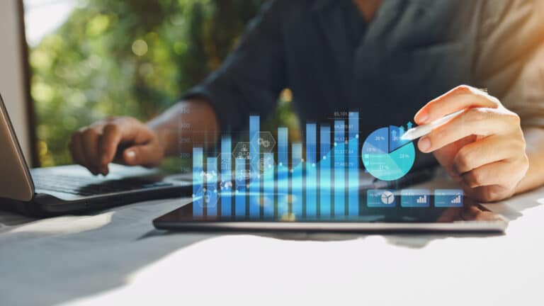 Person using a laptop and tablet with holographic financial charts and graphs displayed in a bright setting.