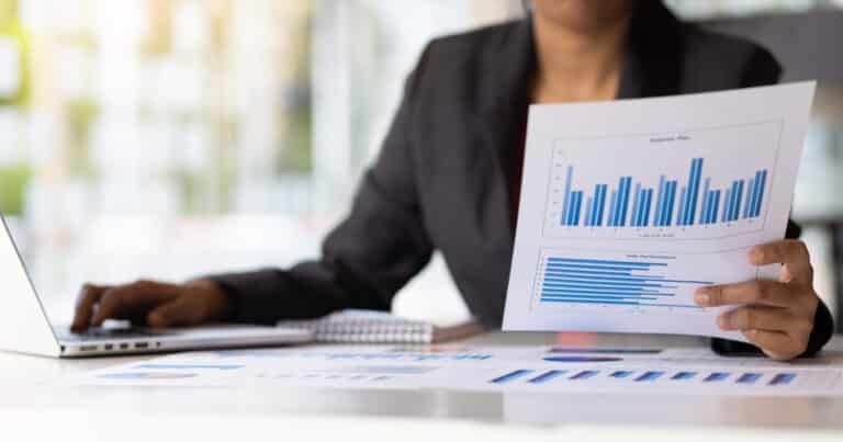 A person in a suit examines bar graphs on paper while using a laptop at a desk with additional papers and a notebook.