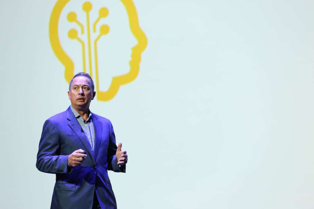 A man in a suit speaks on stage with an abstract brain illustration in the background.