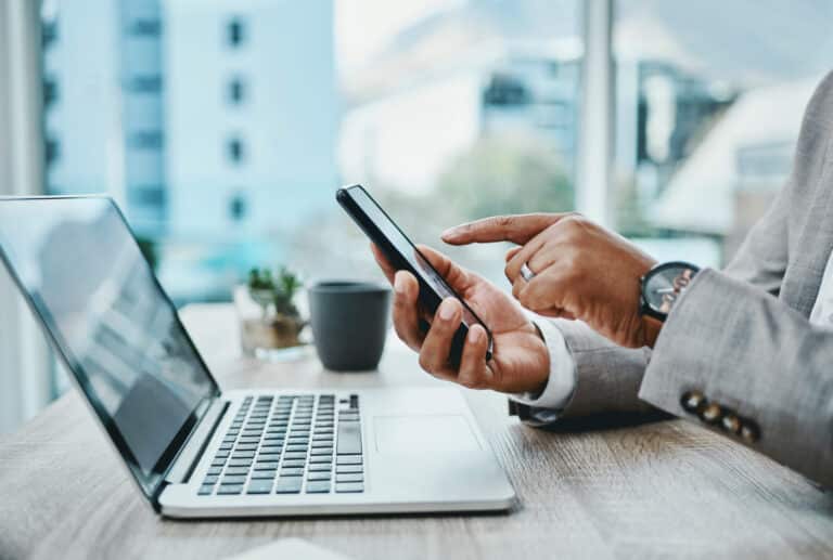 A person in a suit uses a smartphone next to a laptop on a desk.
