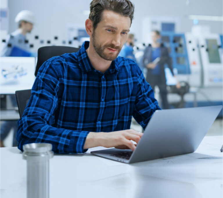 A man in a blue plaid shirt works on a laptop at a white desk. Office equipment and colleagues in the background.