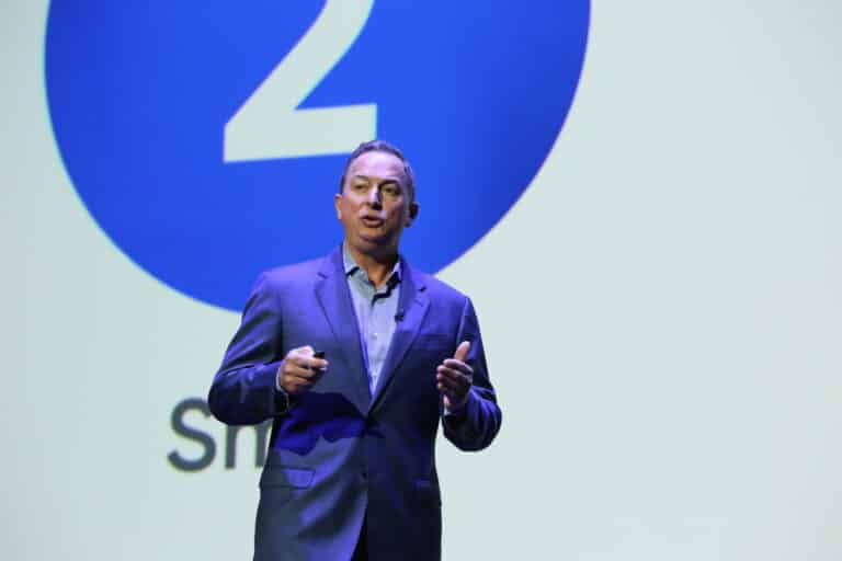 A man in a suit speaks on stage in front of a large screen showing the number 2.