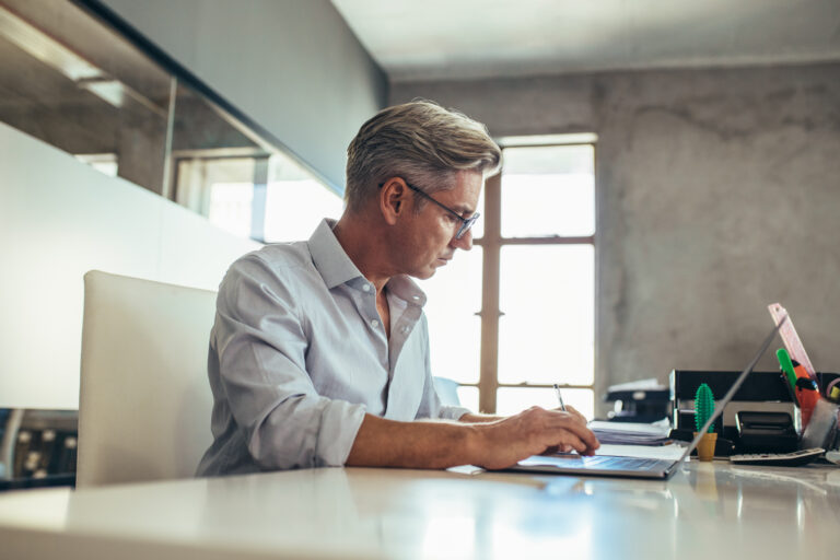Man with gray hair and glasses working intently on a laptop at a desk, with a notebook and office supplies around him.
