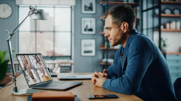 A man in a blue sweater participates in a video call on a laptop in a home office setting.