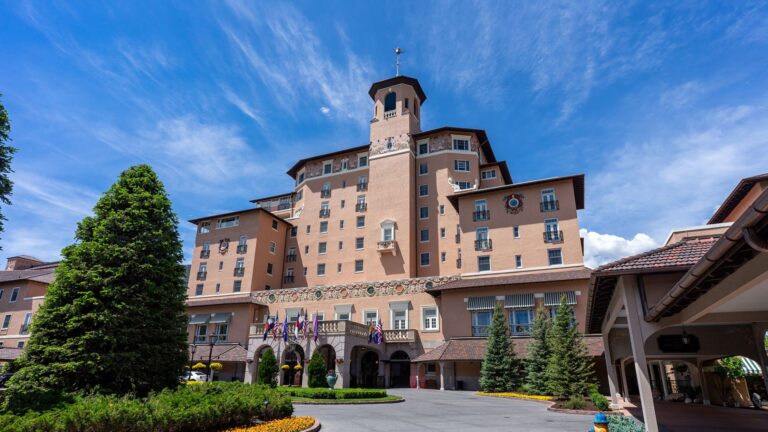 Large, historic hotel with a central tower and ornate exterior, surrounded by trees and flowers under a clear blue sky.