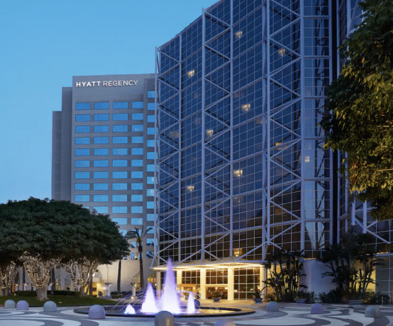 Night view of Hyatt Regency hotel with a modern glass facade and a lit water fountain in the foreground. Trees with string lights are on either side of the entrance.