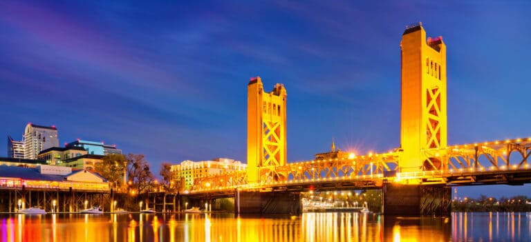 Golden Tower Bridge illuminated at dusk, reflecting on the calm river, with city buildings in the background under a clear sky.
